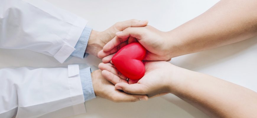 A doctor's hands holding a woman's hand, which cradles a heart-shaped object, indicating potential heart attack symptoms
