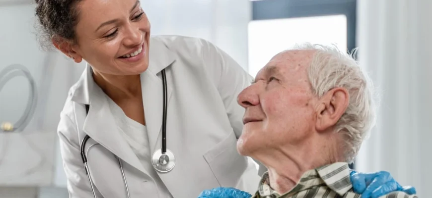 A smiling female doctor with a stethoscope is looking at and talking with an elderly male patient, discussing Eliquis cost or medication. The doctor is gently touching the patient's shoulders.